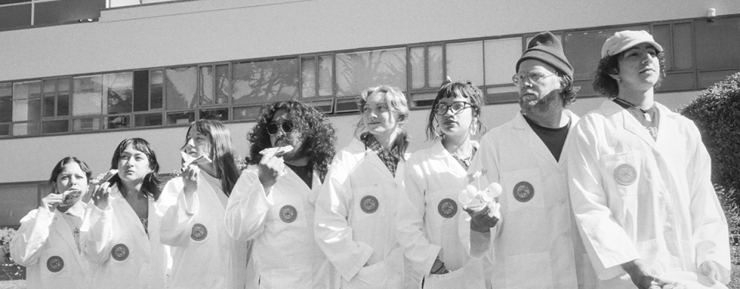 Black and white photo of eight SFSU students in lab coats standing on the Quad with four of them each eating a slice of pizza